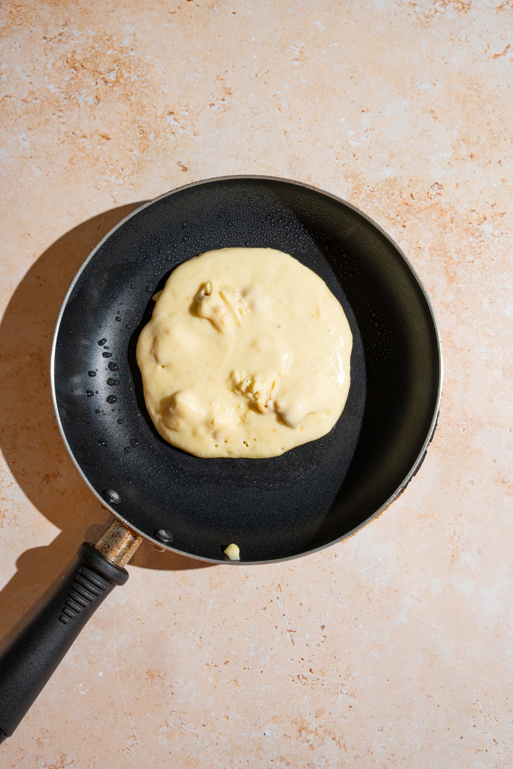 A skillet with an uncooked IHOP New York cheesecake pancake. The skillet is on a tan counter.