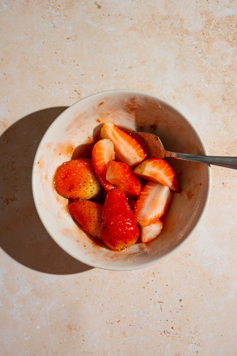 A bowl with a spoon mixing a strawberry topping mixture. The bowl is on a tan counter.