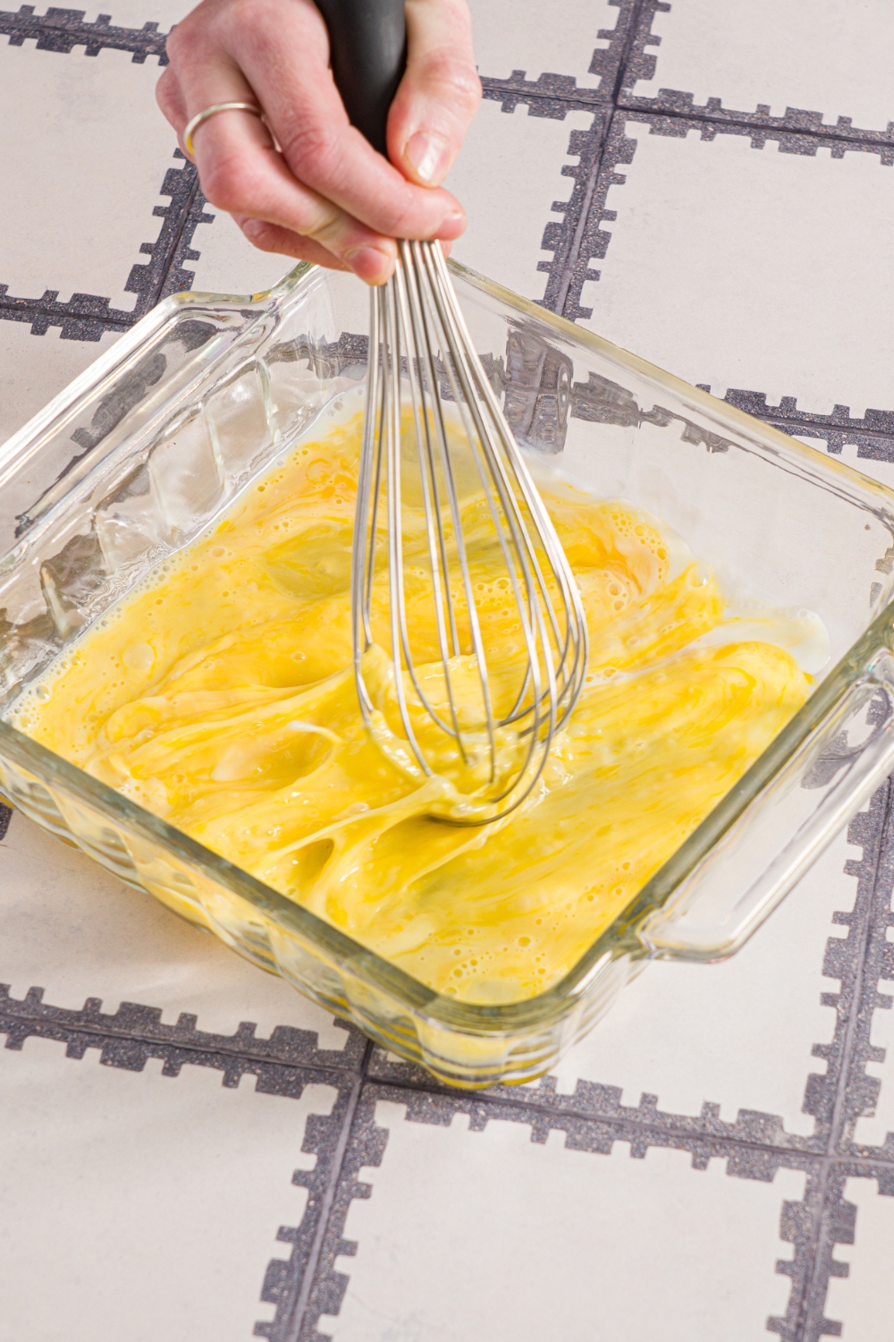 A glass baking dish with a whisk mixing eggs and milk. The dish is on a tiled counter.
