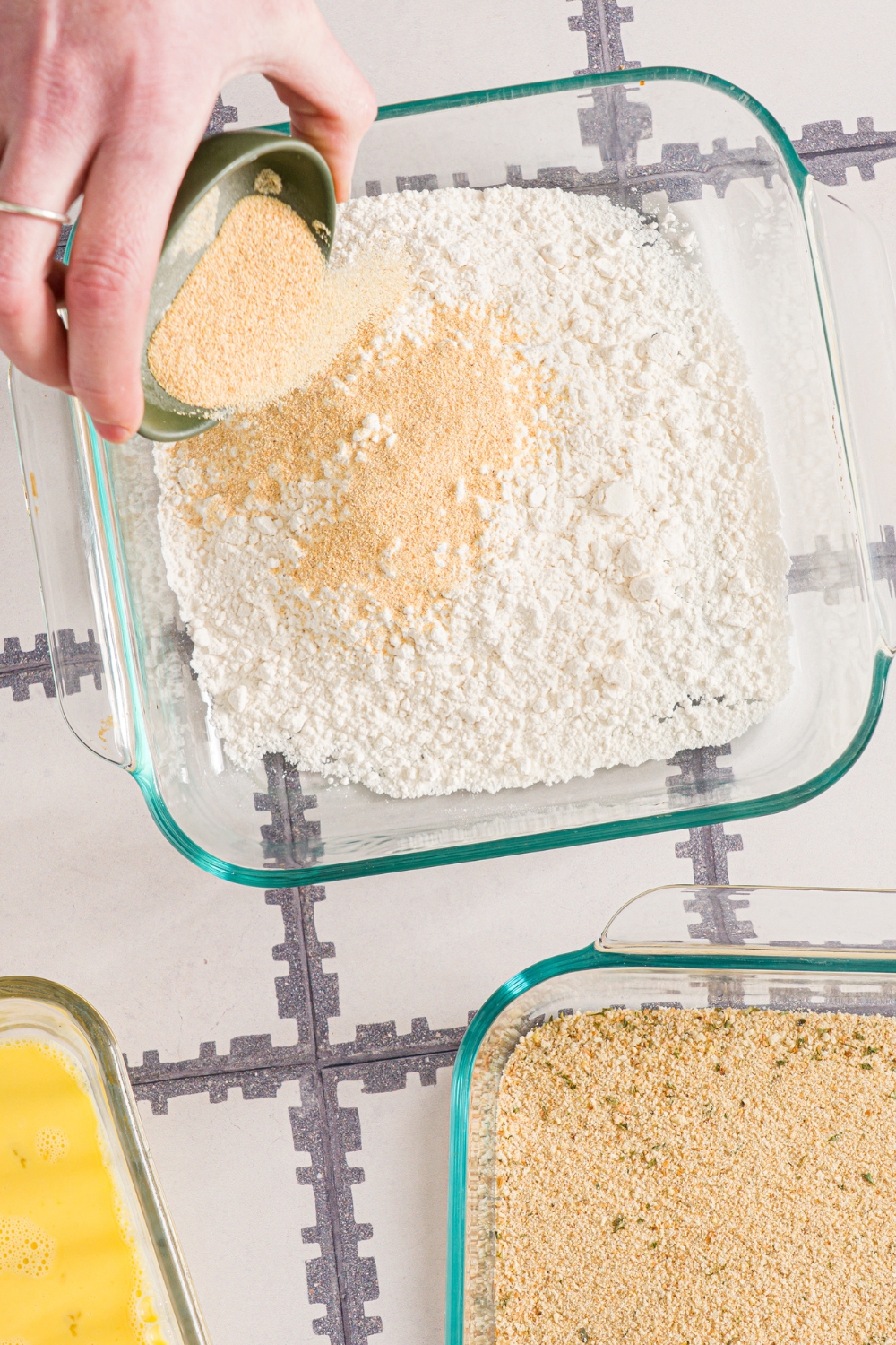A glass baking dish with flour with a small bowl of seasoning being dumped into the dish. The dish is on a tiled counter.