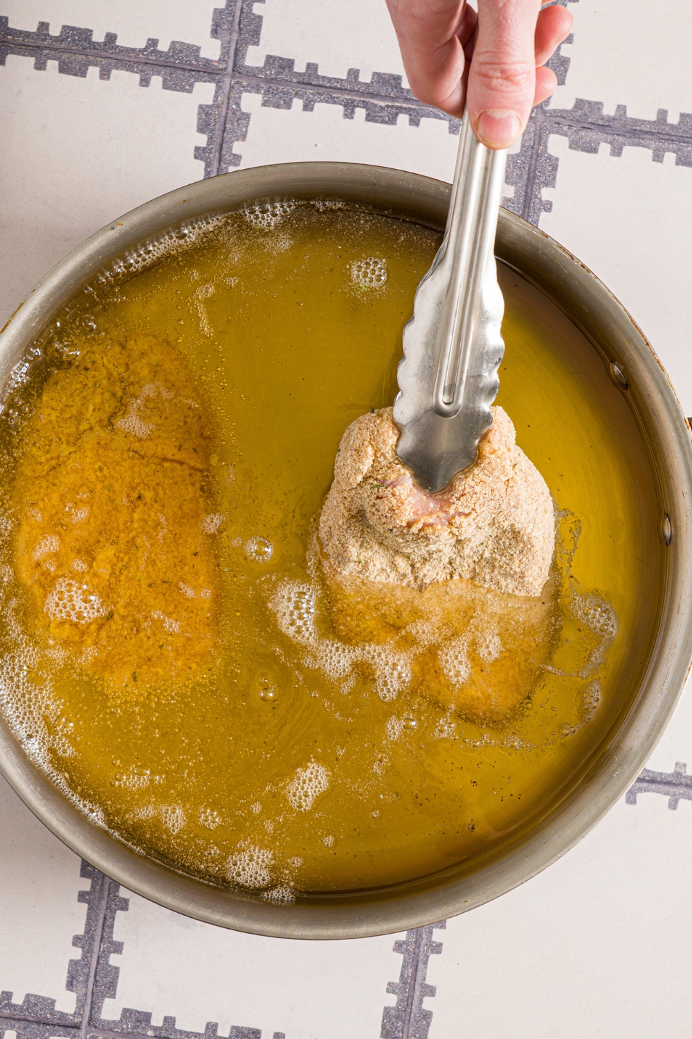 A skillet with breaded pork cutlets being fried in oil. The skillet is on a tiled counter.