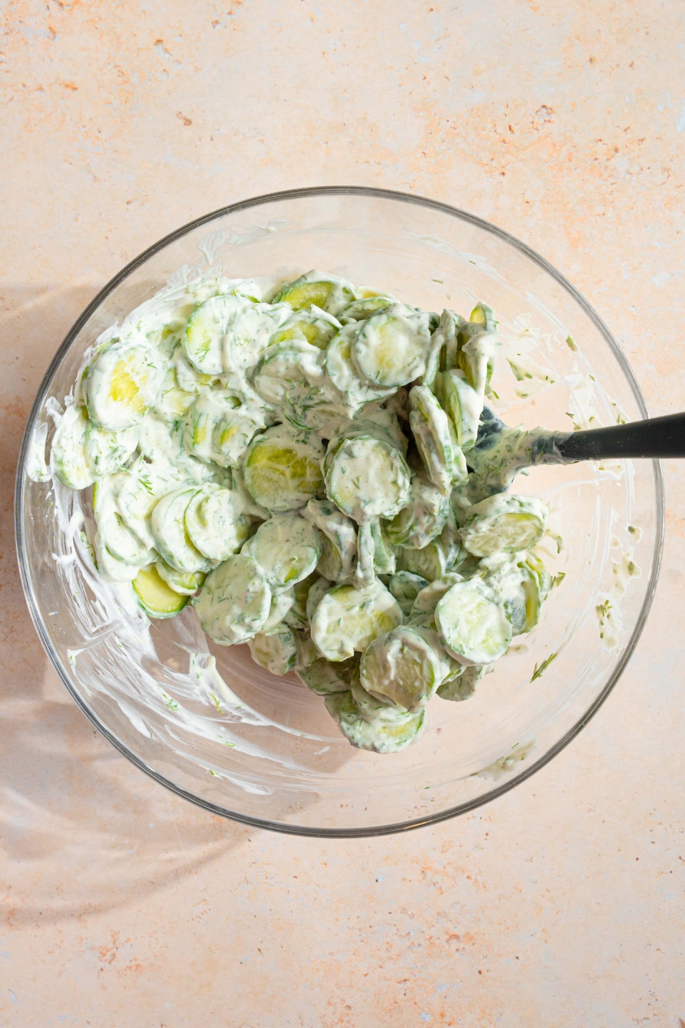 A glass bowl with a spatula mixing sliced cucumbers with a cucumber dill dressing. The bowl is on a tan counter.