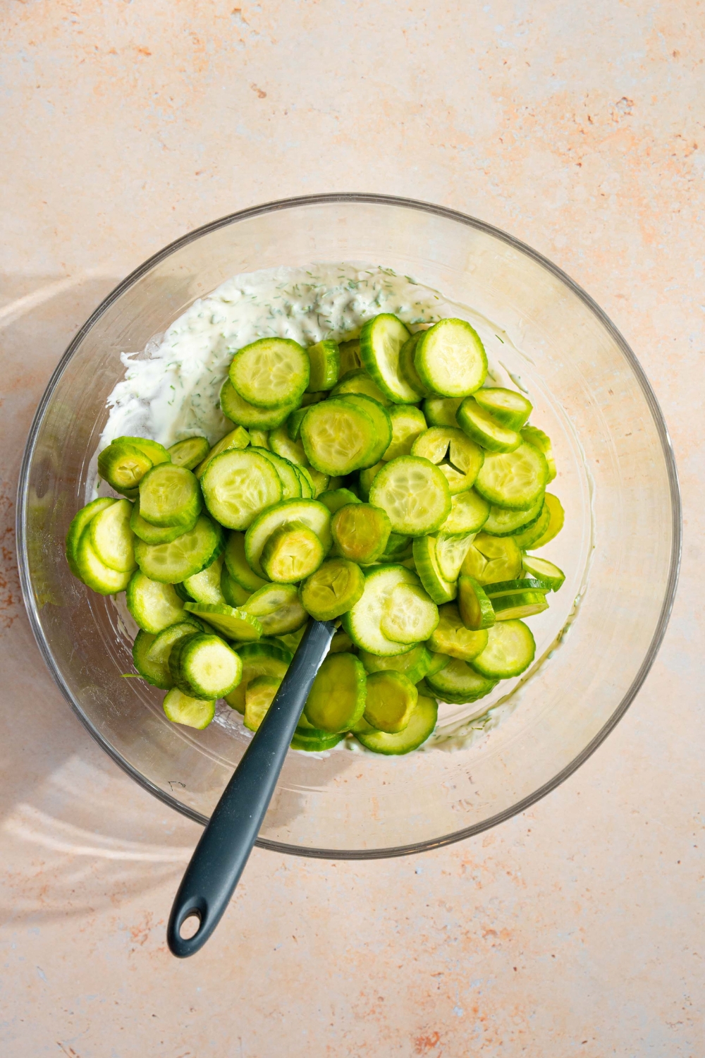 A glass bowl with sliced cucumber on top of a cucumber dill dressing. There is a spatula in the bowl. The bowl is on a tan counter.