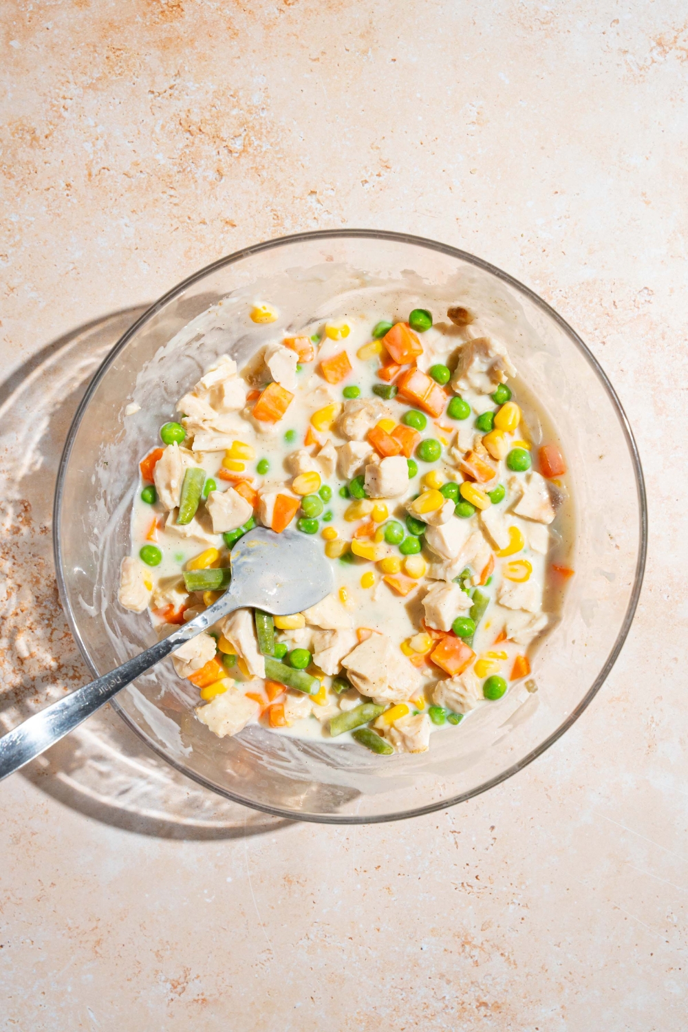 A glass bowl with a spoon stirring a chicken pot pie mixture including cream of chicken soup, cubed chicken, frozen mixed vegetables, and seasonings. The bowl is on a tan counter.
