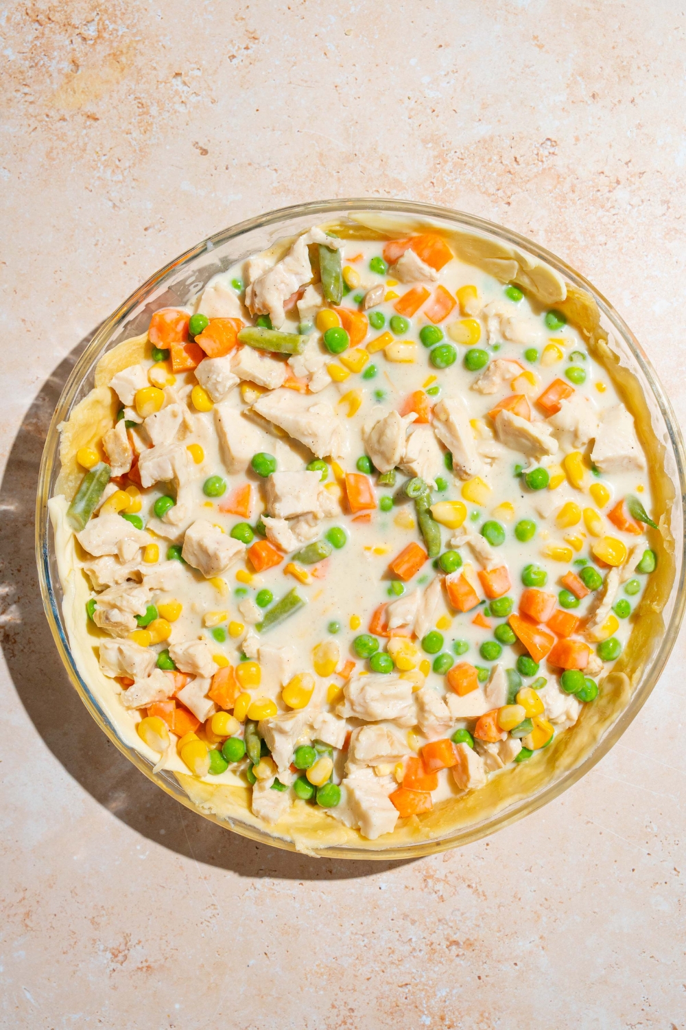 An open pie dish with chicken pot pie filling on a bottom pie crust. The dish is on a tan counter.