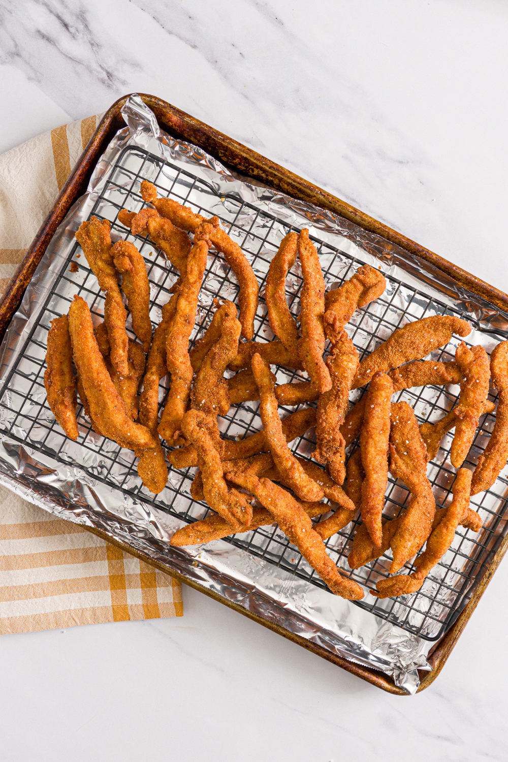 A baking sheet lined with foil and with a rack with fried chicken fries seasoned with salt. The sheet is on a marble counter with a yellow checkered napkin.