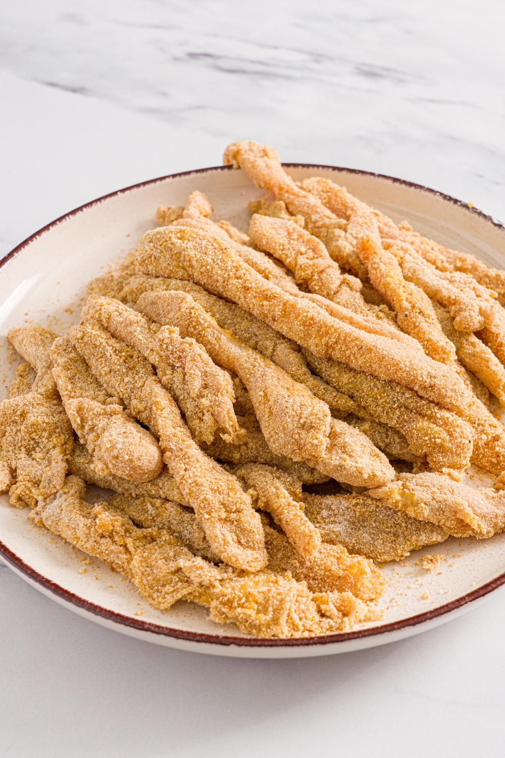 A plate with breaded uncooked chicken fries. The plate is on a marble counter.