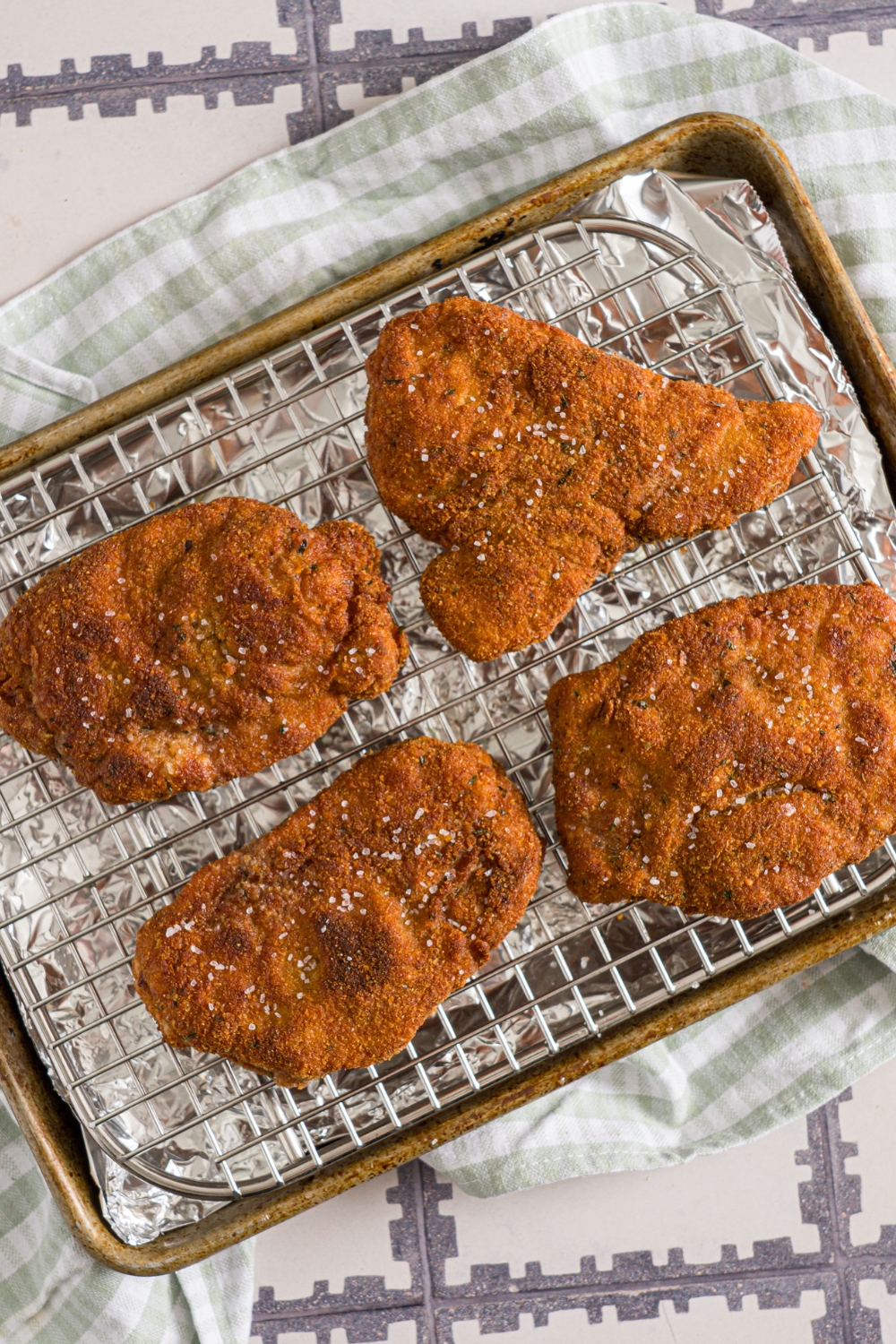 A baking sheet lined with foil and a rack with four pieces of crispy breaded pork cutlets seasoned with salt. The sheet is on a green cloth napkin on a tiled counter.