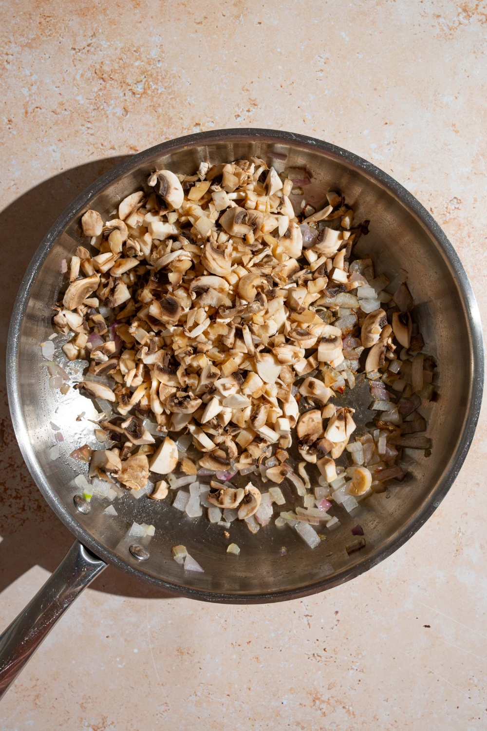 A skillet with diced mushrooms and onions cooking in oil. The skillet is on a tan counter.