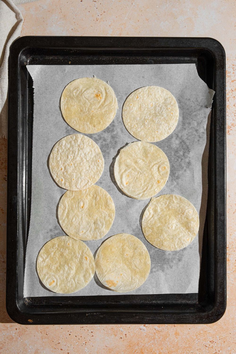 A baking sheet lined with parchment paper with several mini tortilla shells. The sheet is on a tan counter.