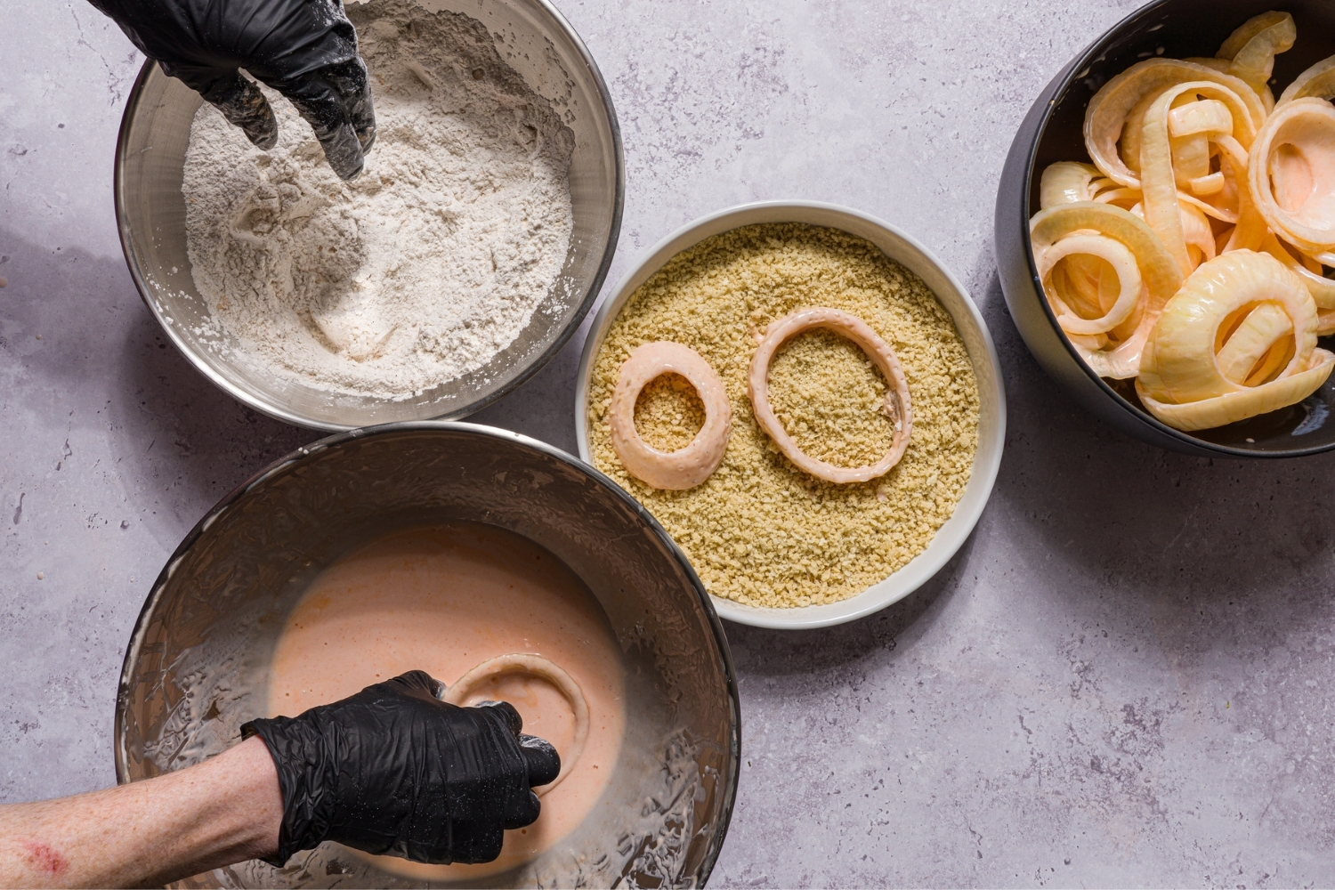 Several bowls with ingredients to bread onion rings including a flour mixture, breadcrumbs, a buttermilk mixture, and a bowl of buttermilk soaked onion rings. Onion rings are being dredged in the bowls.
