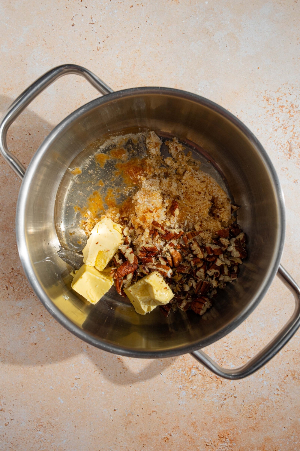 A stock pot with ingredients to make pecan pie topping including pecans, corn syrup, butter, brown sugar, vanilla, and egg. The pot is on a tan counter.
