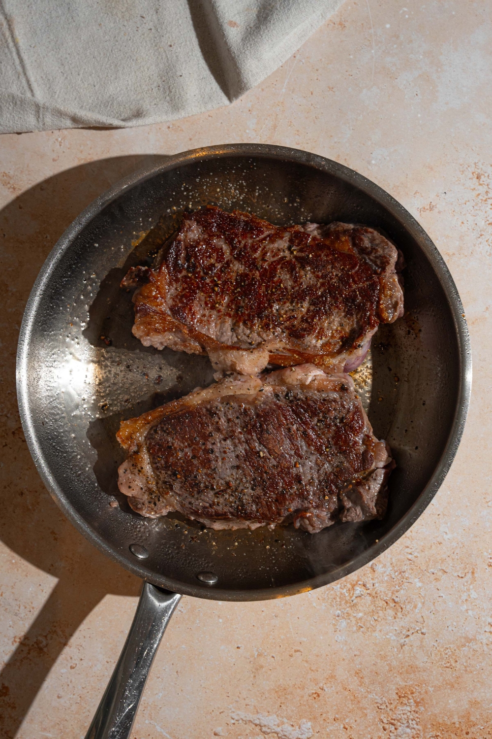 A skillet with two steaks searing in oil. The skillet is on a tan counter.