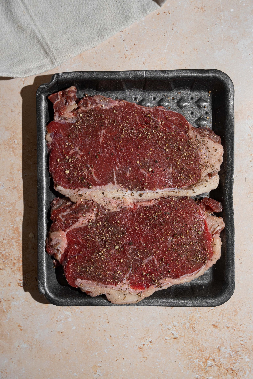 Two uncooked steaks seasoned with salt and pepper. The steaks are on a tan counter.