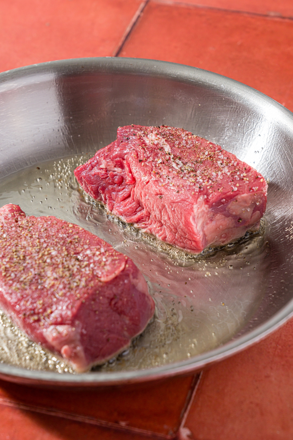 A stainless steel skillet with two uncooked petite sirloin steaks searing in oil. The skillet is on a red tiled counter.