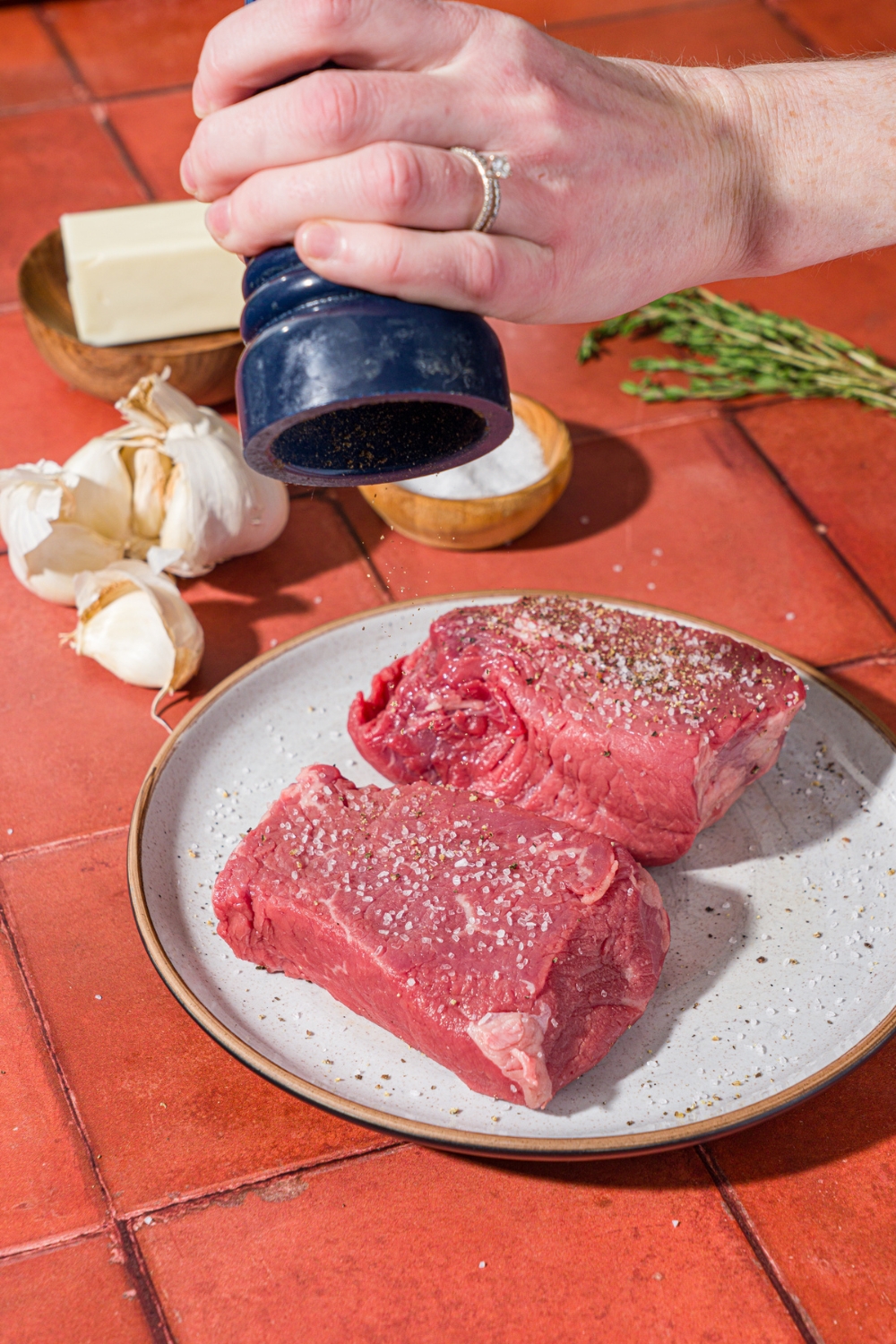 A ceramic plate with two uncooked petite sirloin steaks seasoned with salt. A hand is using a pepper mill to season the steaks. The plate is on a red tiled counter with garlic, rosemary, and a small bowl of salt.