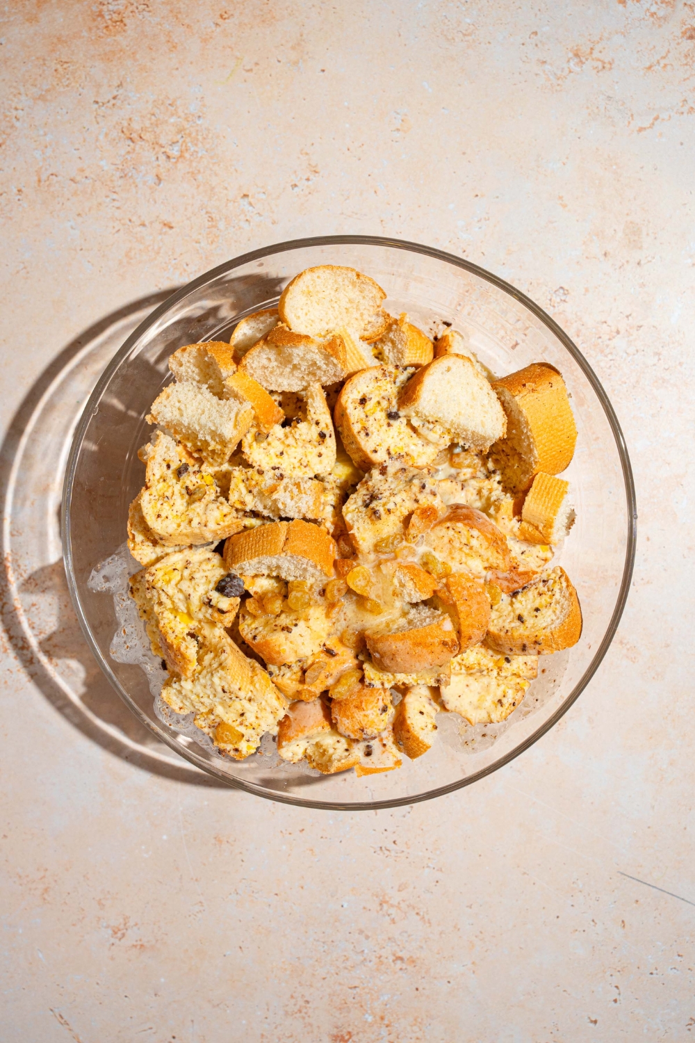A glass bowl with cubed bread topped with a custard mixture. The bowl is on a tan counter.