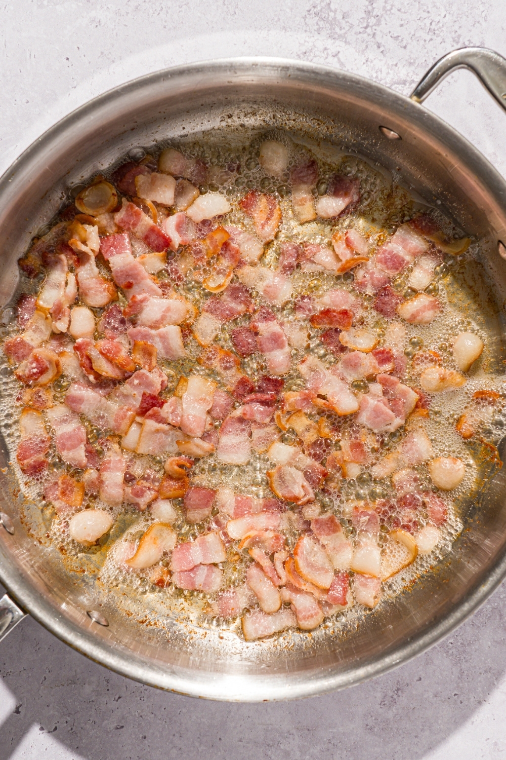 A skillet with sizzling chopped bacon. The skillet is on a white counter.