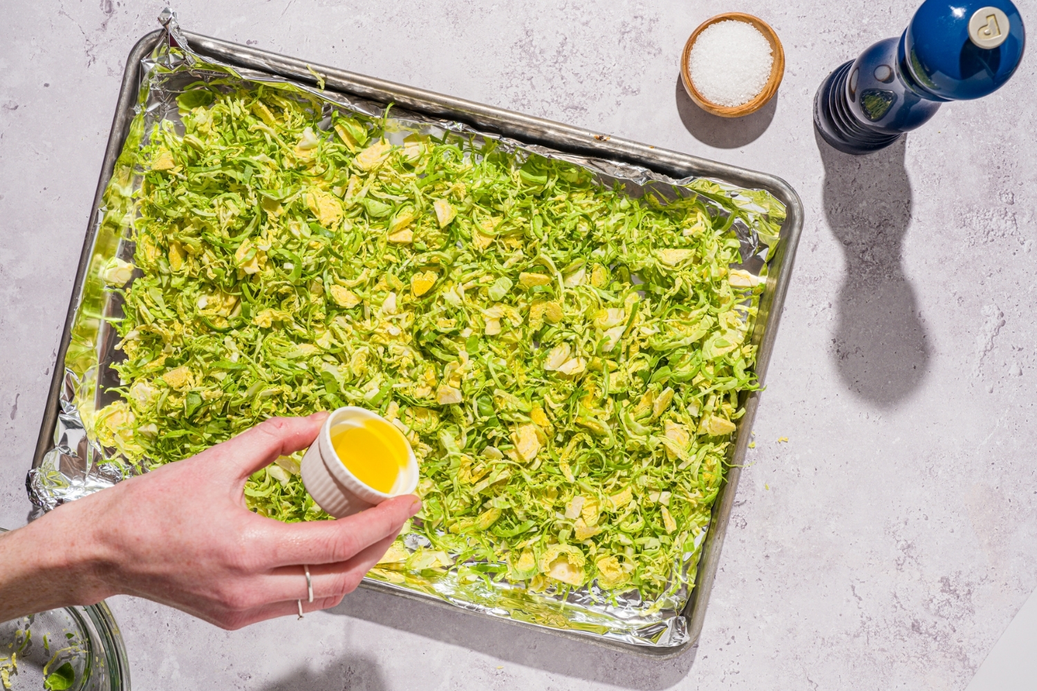 A baking sheet lined with foil with shaved brussel sprouts. A hand is pouring oil from a small ramekin onto the sprouts. The sheet is on a white counter with a small bowl of salt and pepper mill.
