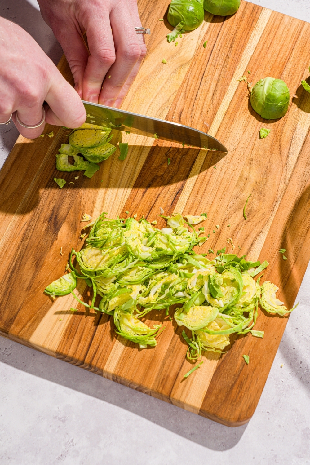 A hand trimming and shaving brussel sprouts with a knife on a wooden board. The board is on a white counter.
