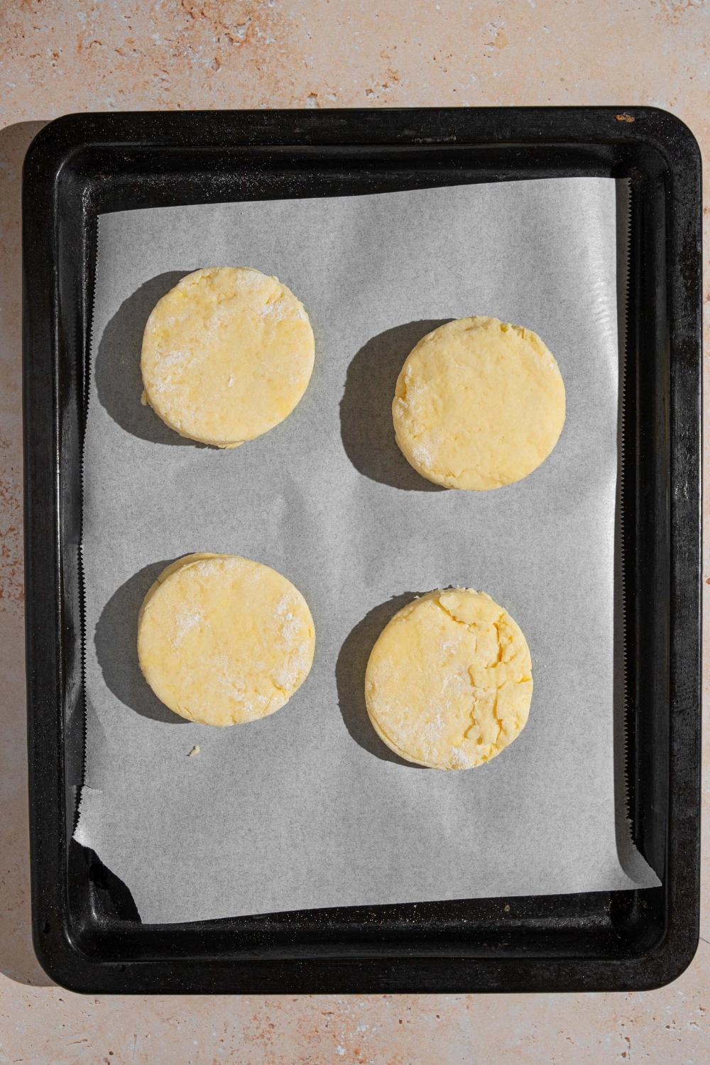 A baking sheet lined with parchment paper with four cuts of uncooked shortcake dough. The sheet is on a tan counter.