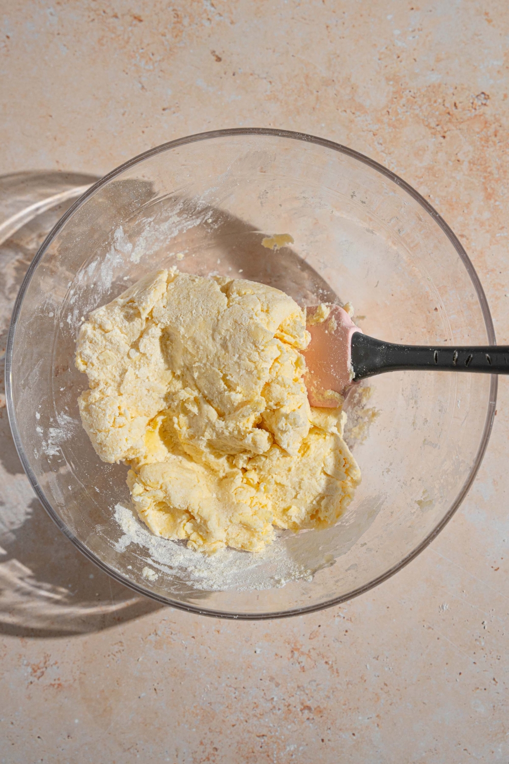A glass bowl with a spatula mixing half and half with shortcake dough. The bowl is on a tan counter.