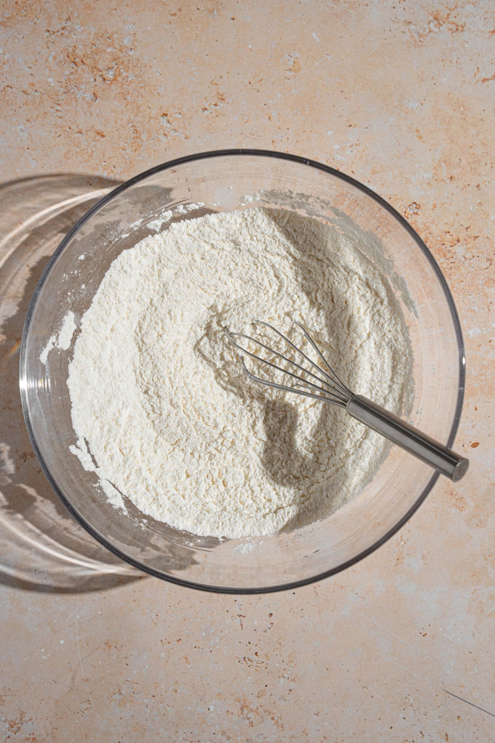 A glass bowl with a whisk mixing dry ingredients to make shortcake dough. The bowl is on a tan counter.