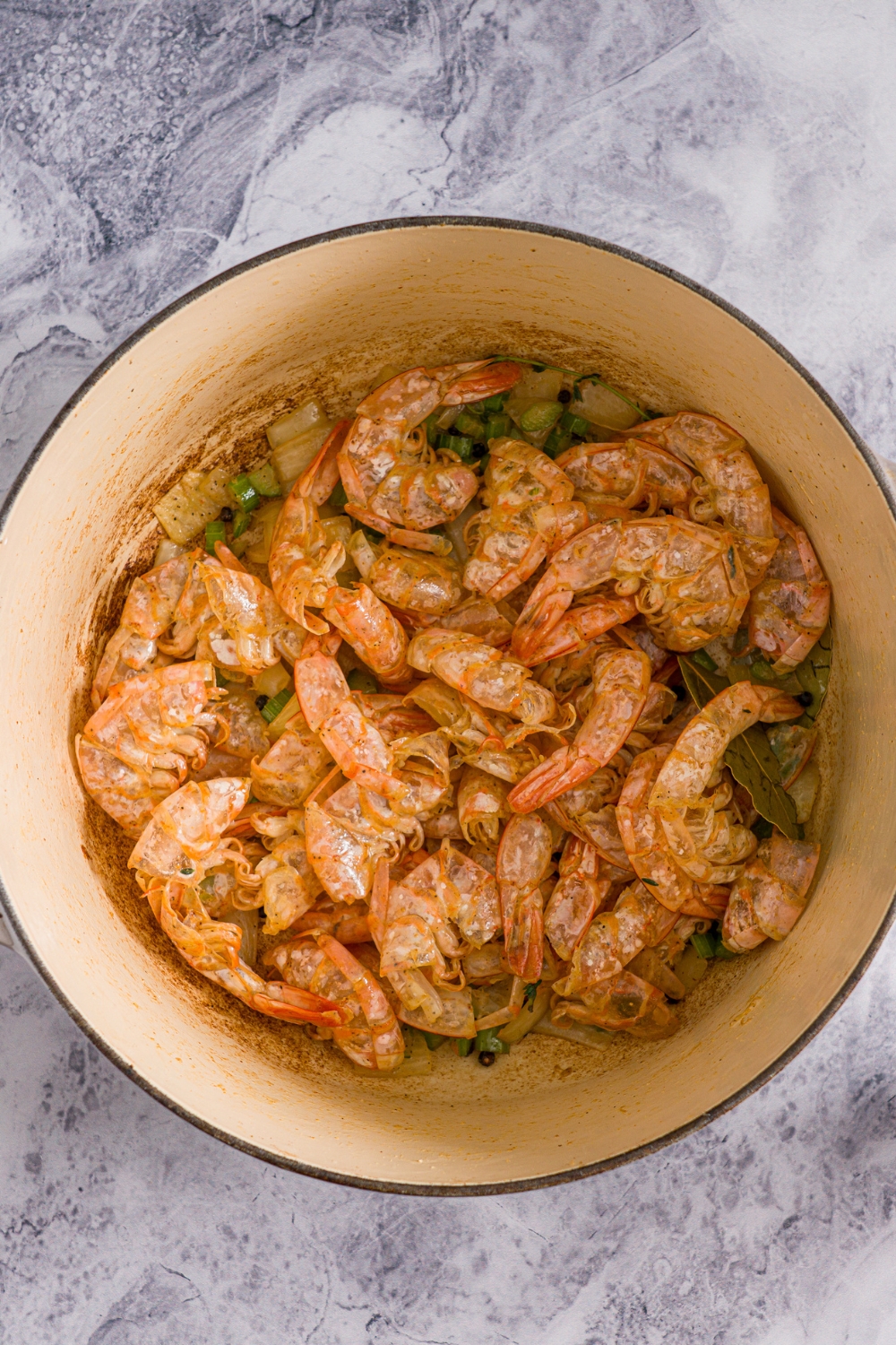 A dutch oven with shrimp shells cooked in butter, oil, diced vegetables, and bay leaves. The pot is on marble counter.