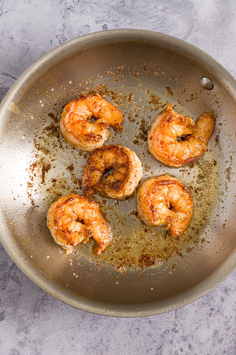 A skillet with shrimp sautéing in oil. The skillet is on a marble counter.