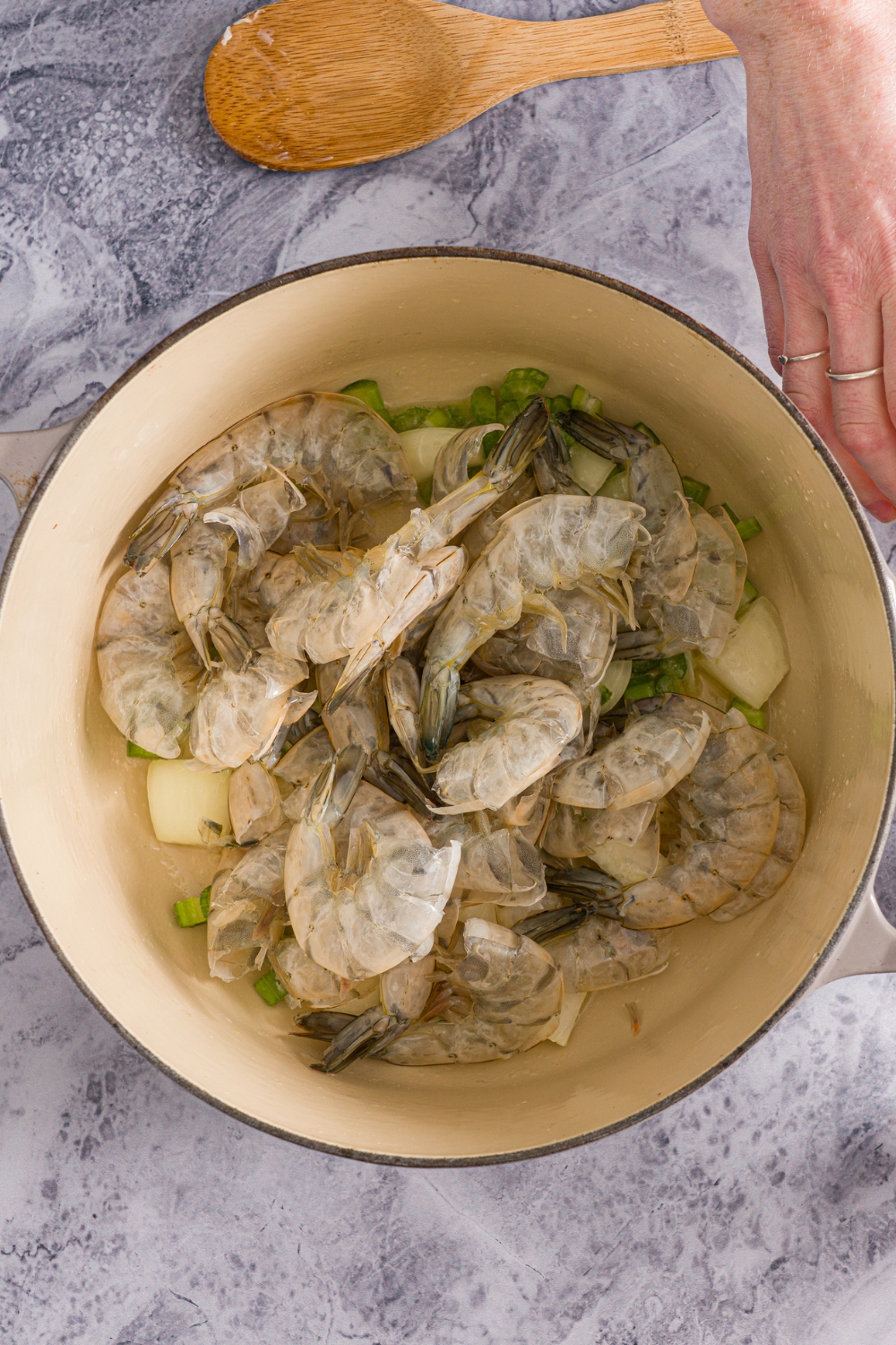 A dutch oven with peeled shrimp shells with diced celery and onion. The pot is on a marble counter.