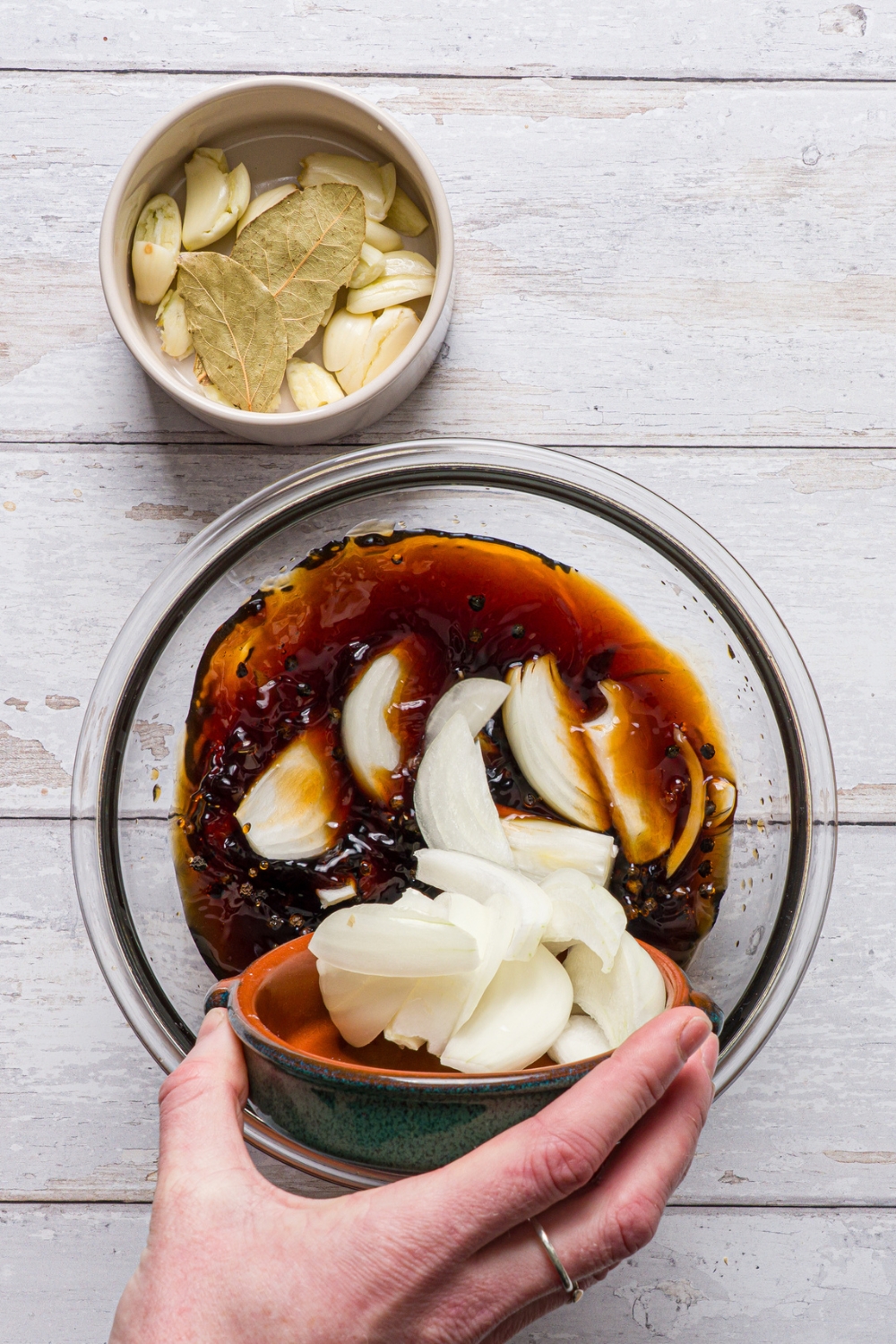 A glass bowl with ingredients to make adobo sauce including sliced white onion, soy sauce, brown sugar, and red wine vinegar. A hand is pouring sliced white onion into the bowl. The bowl is on a wood counter with a small bowl of garlic cloves and bay leaves.