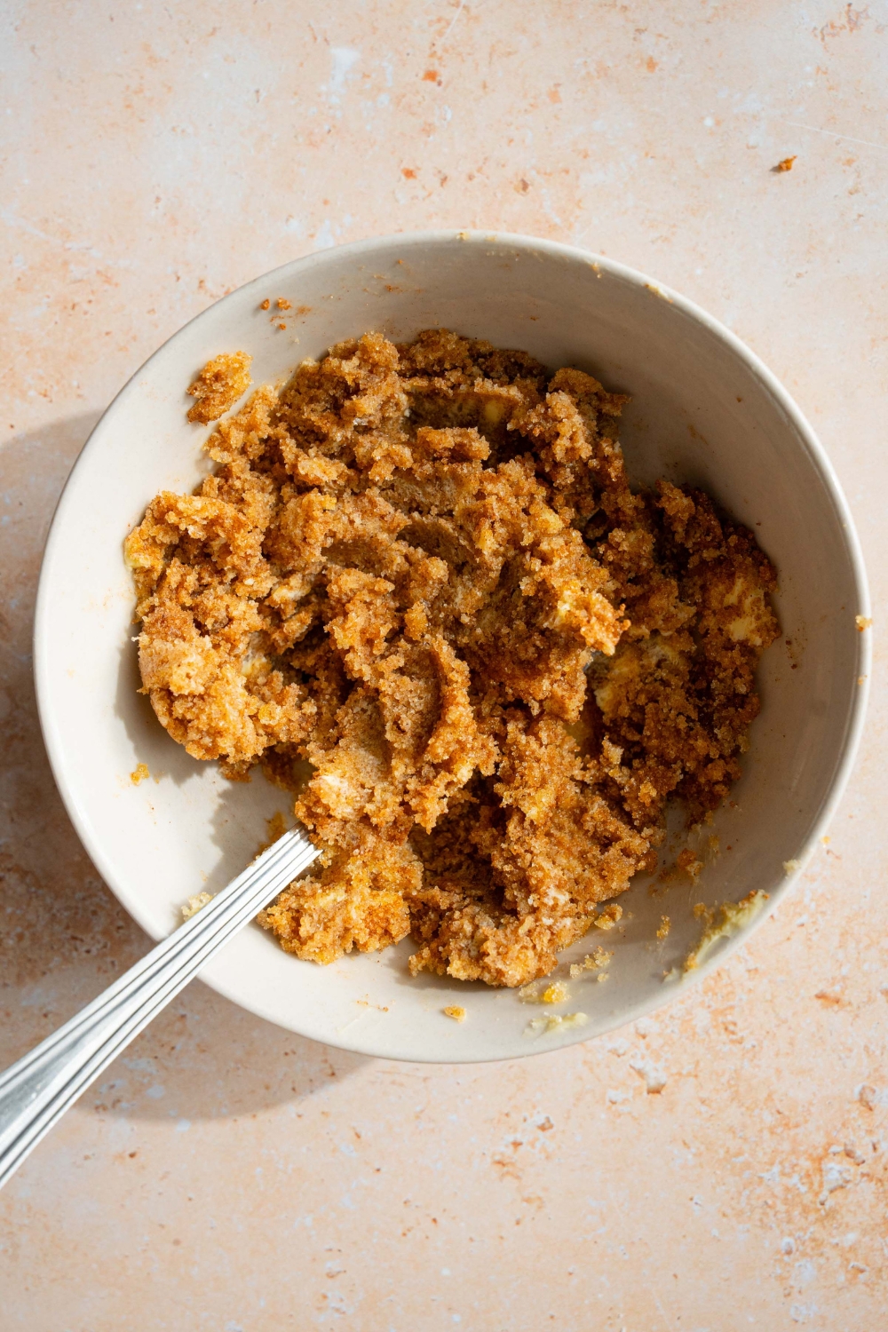 A large white bowl with a fork mixing a cinnamon sugar mixture including cinnamon, butter, and brown sugar. The bowl is on a tan counter.