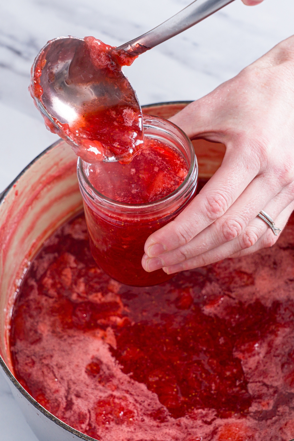 A dutch oven with cooked strawberry jam with a ladle pouring jam into a can.