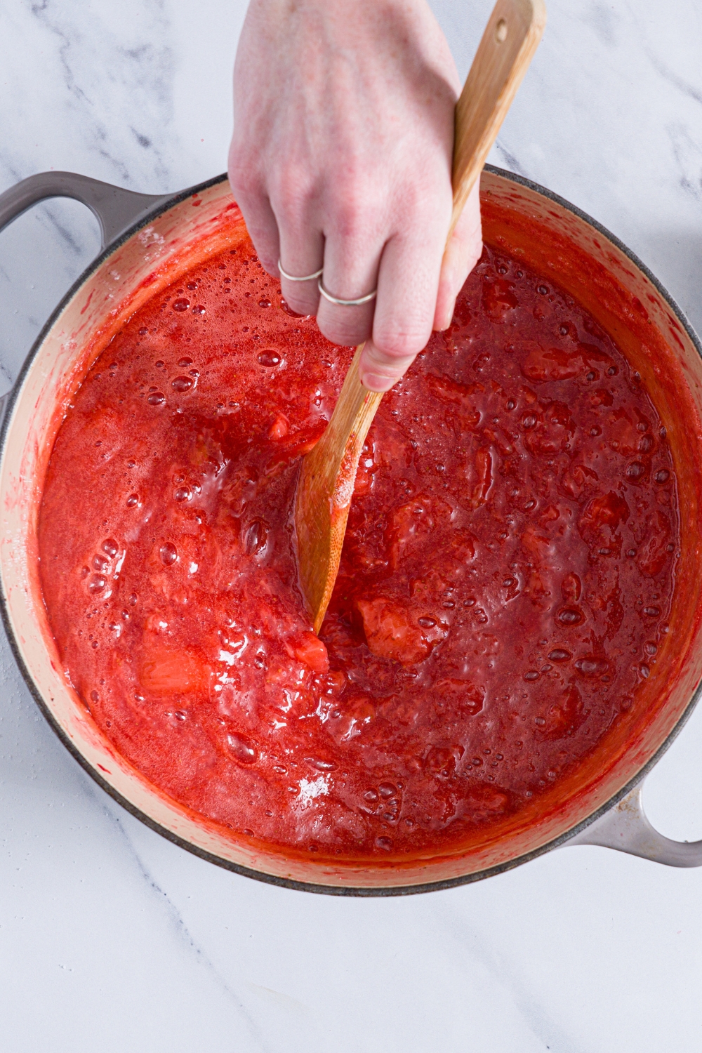 A dutch oven with boiling strawberry jam and a wooden spoon stirring the jam. The oven is on a marble counter.