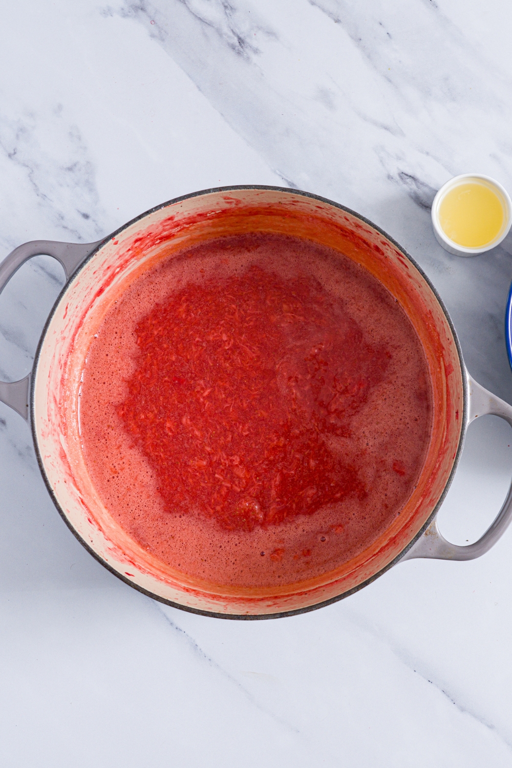 A dutch oven with cooked strawberry jam on a marble counter with a small bowl of lemon juice.