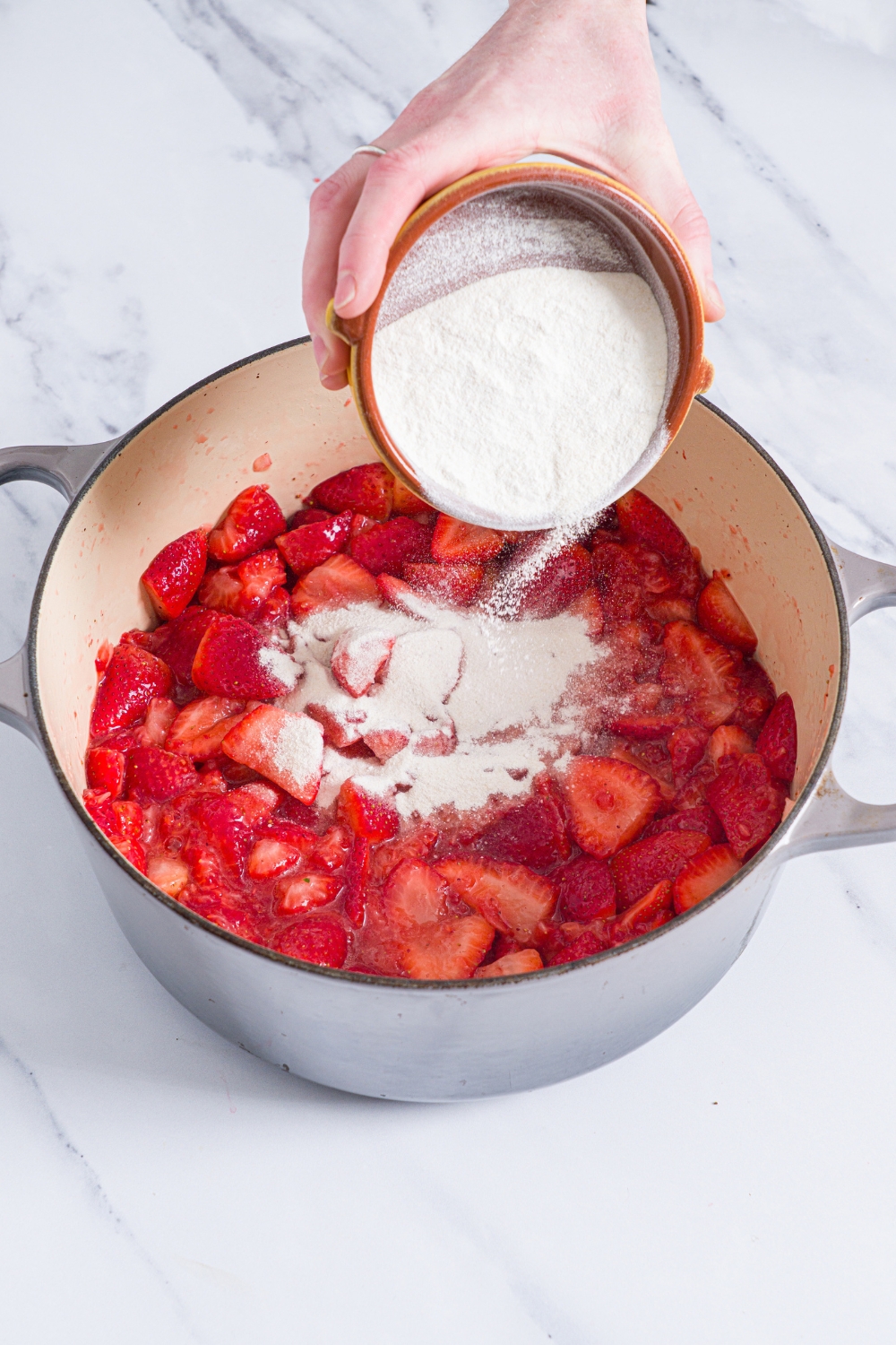 A dutch oven with crushed strawberries with a bowl of pectin being poured on the strawberries. The pot is on a marble counter.