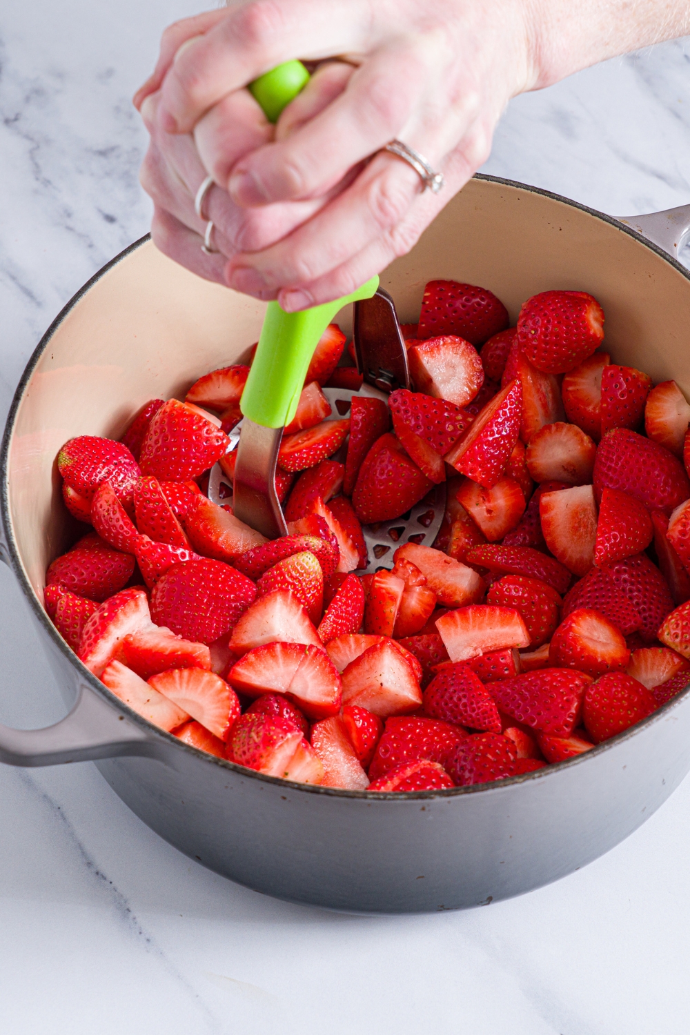 A dutch oven with washed and hulled strawberries. There is a masher crushing the strawberries. The pot is on a marble counter.
