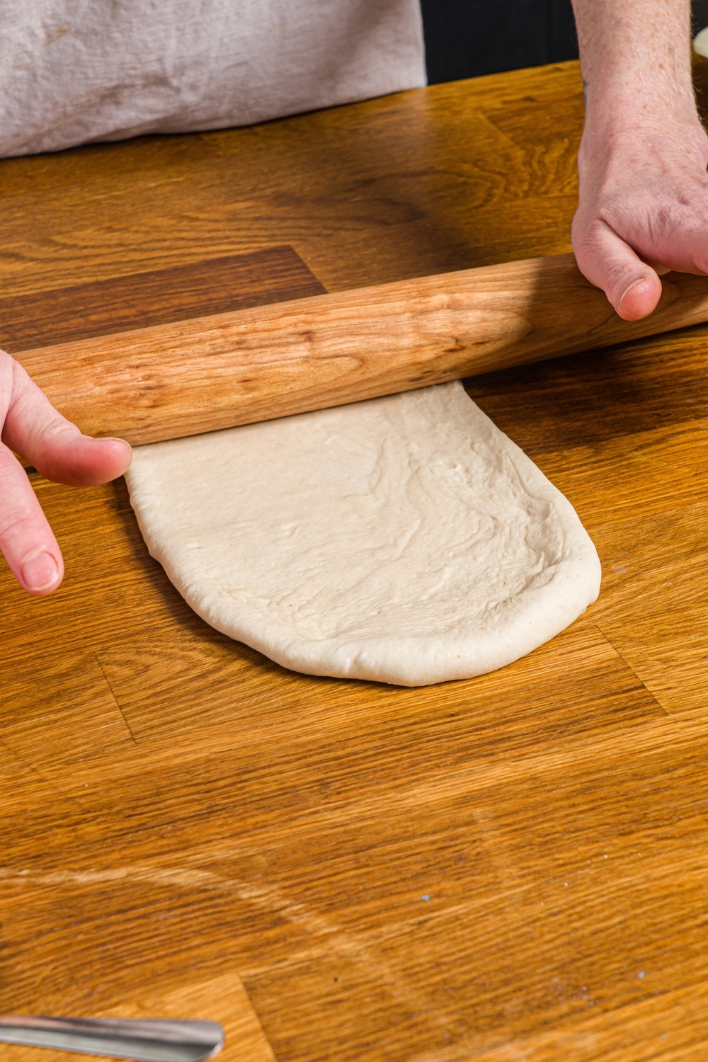 A pair of hands rolling out pizza dough with a wooden roller. The dough is on a wood counter.