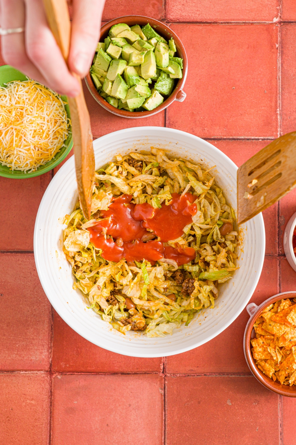 A white bowl with a taco salad mixture topped with catalina dressing. Two salad forks are tossing the salad. The bowl is on a red tiled counter with small bowls of shredded mexican cheese, avocado, and doritos.