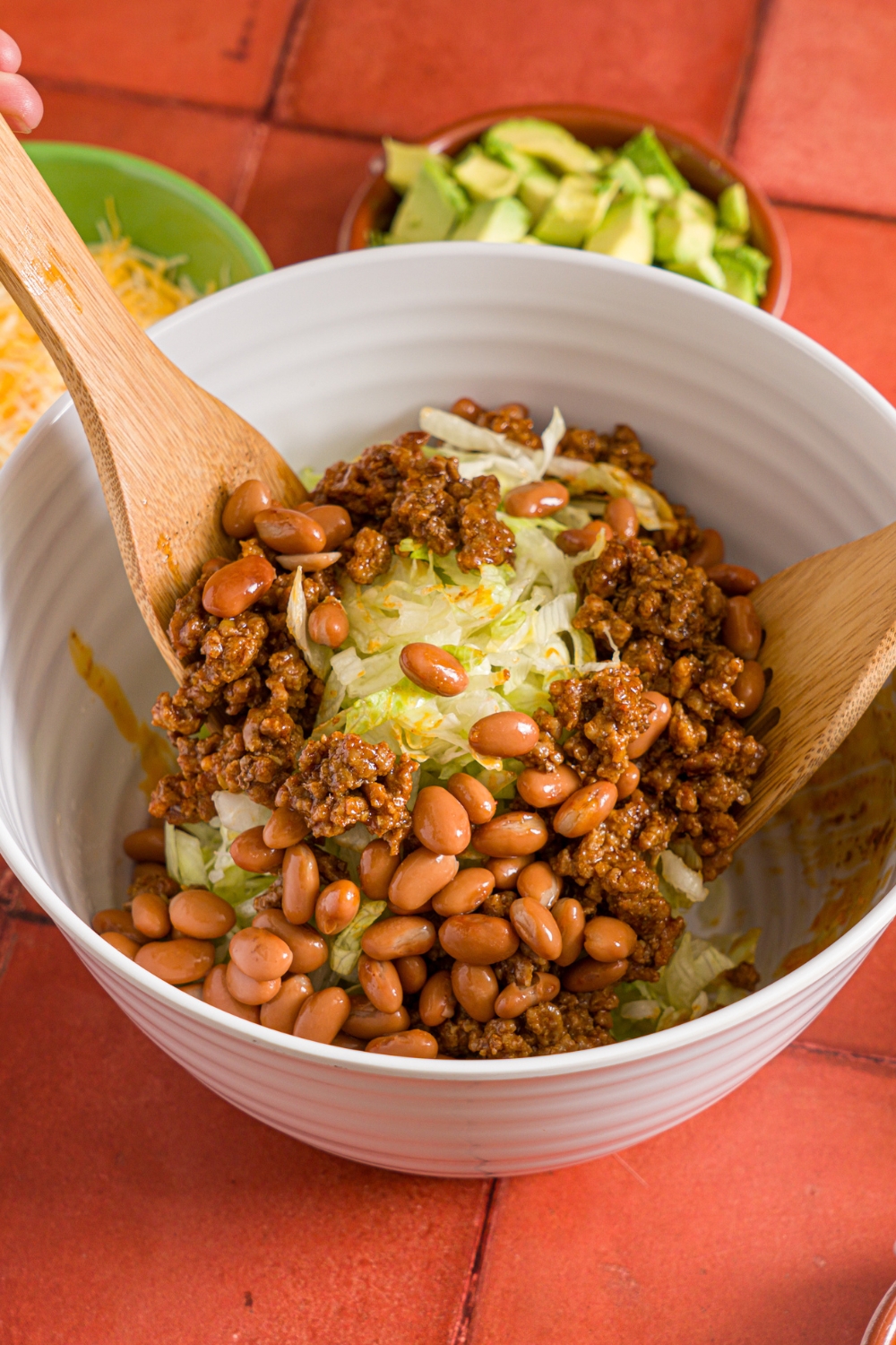 A bowl of two salad forks mixing a salad mixture including shredded lettuce, chili beans, and taco seasoned ground beef. The bowl is on a red tiled counter with a small bowl of avocao and shredded cheese.