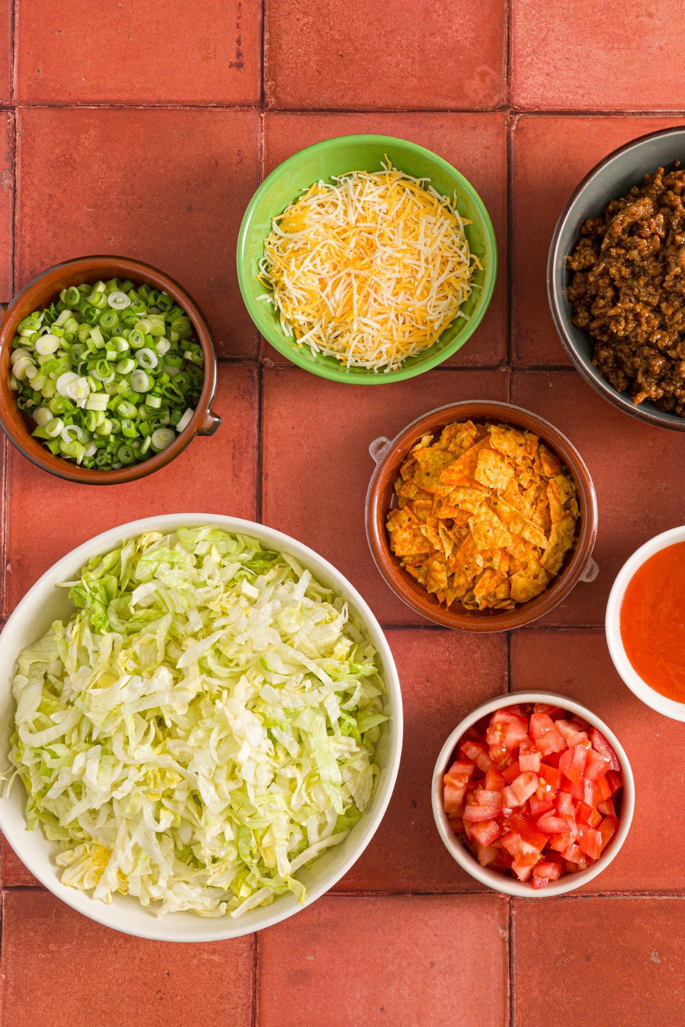 A red tiled counter with several small bowls of taco salad ingredients including shredded lettuce, shredded Mexican cheese, doritos, diced tomatoes, diced green onion, and taco seasoned ground beef.