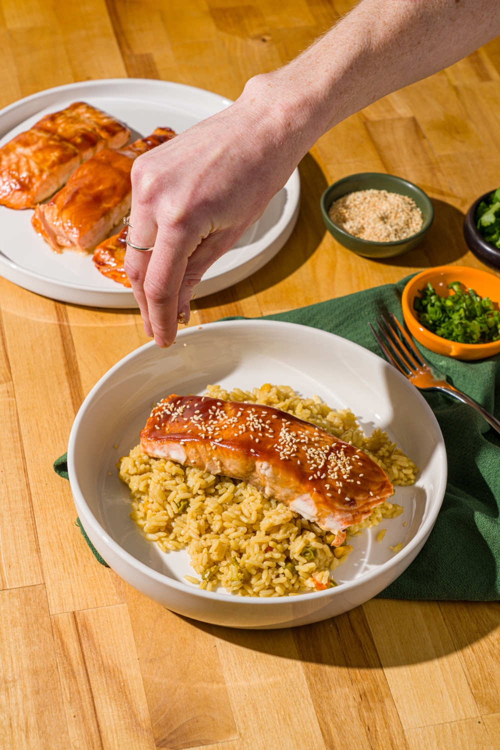 A white bowl with baked teriyaki salmon over fried rice. A hand is sprinkling the salmon with sesame seeds. The bowl is on a wood counter with a plate of baked salmon and green cloth napkin.