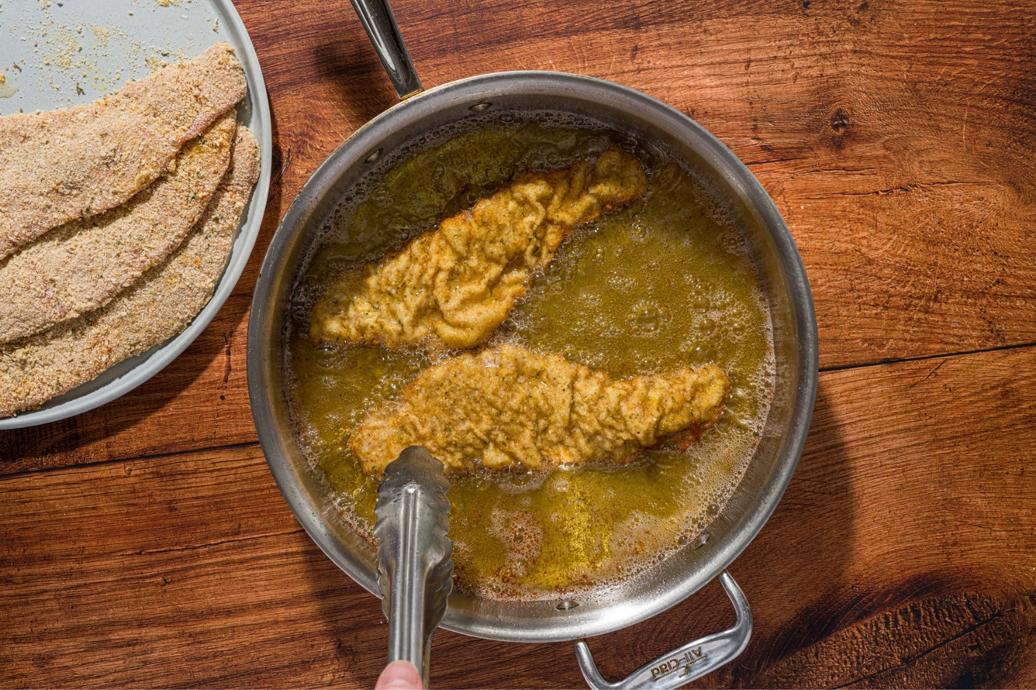 A pot with two breaded veal cutlets frying in oil. A pair of tongs are flipping the cutlets. The pot is on a wood counter with a plate of breaded cutlets.