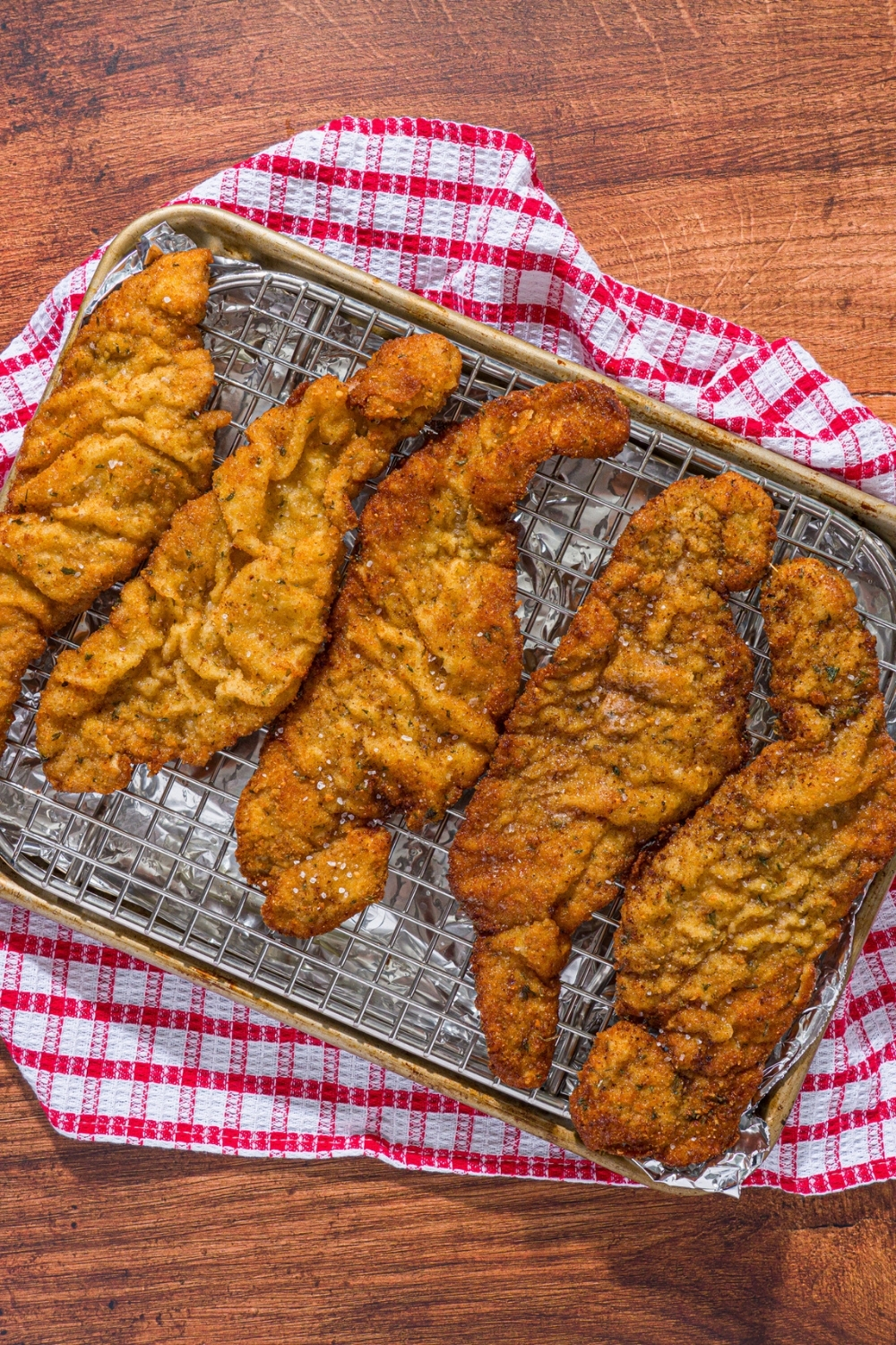 A baking sheet lined with a rack with several pieces of fried breaded veal cutlets. The sheet is on a red checkered napkin on a wood counter.