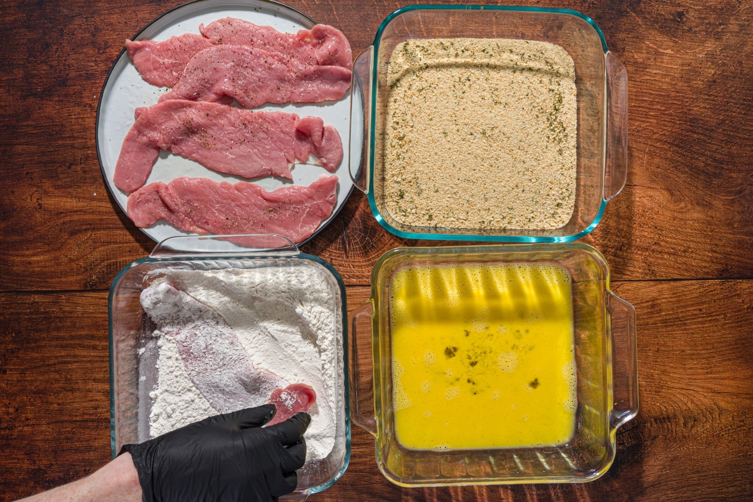 A plate of uncooked veal cutlets with three glass baking dishes with breadcrumbs, flour, and whisked egg.