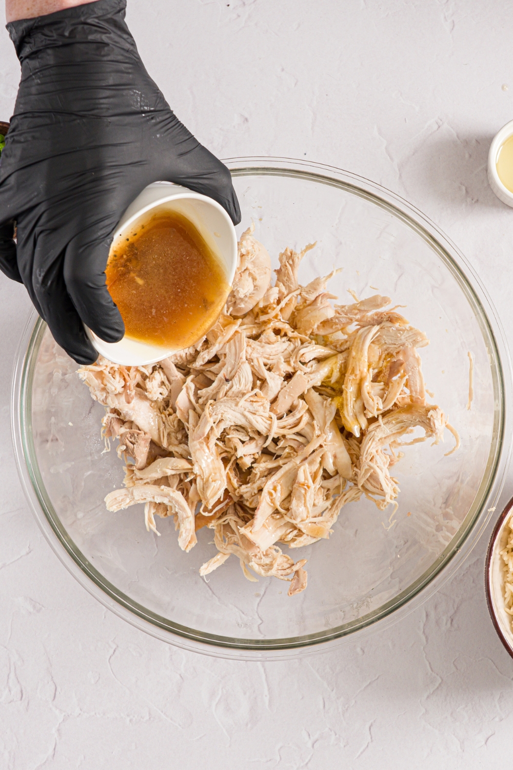 A glass bowl with shredded chicken and a small bowl of sauce being added to the bowl. The bowl is on a white counter.