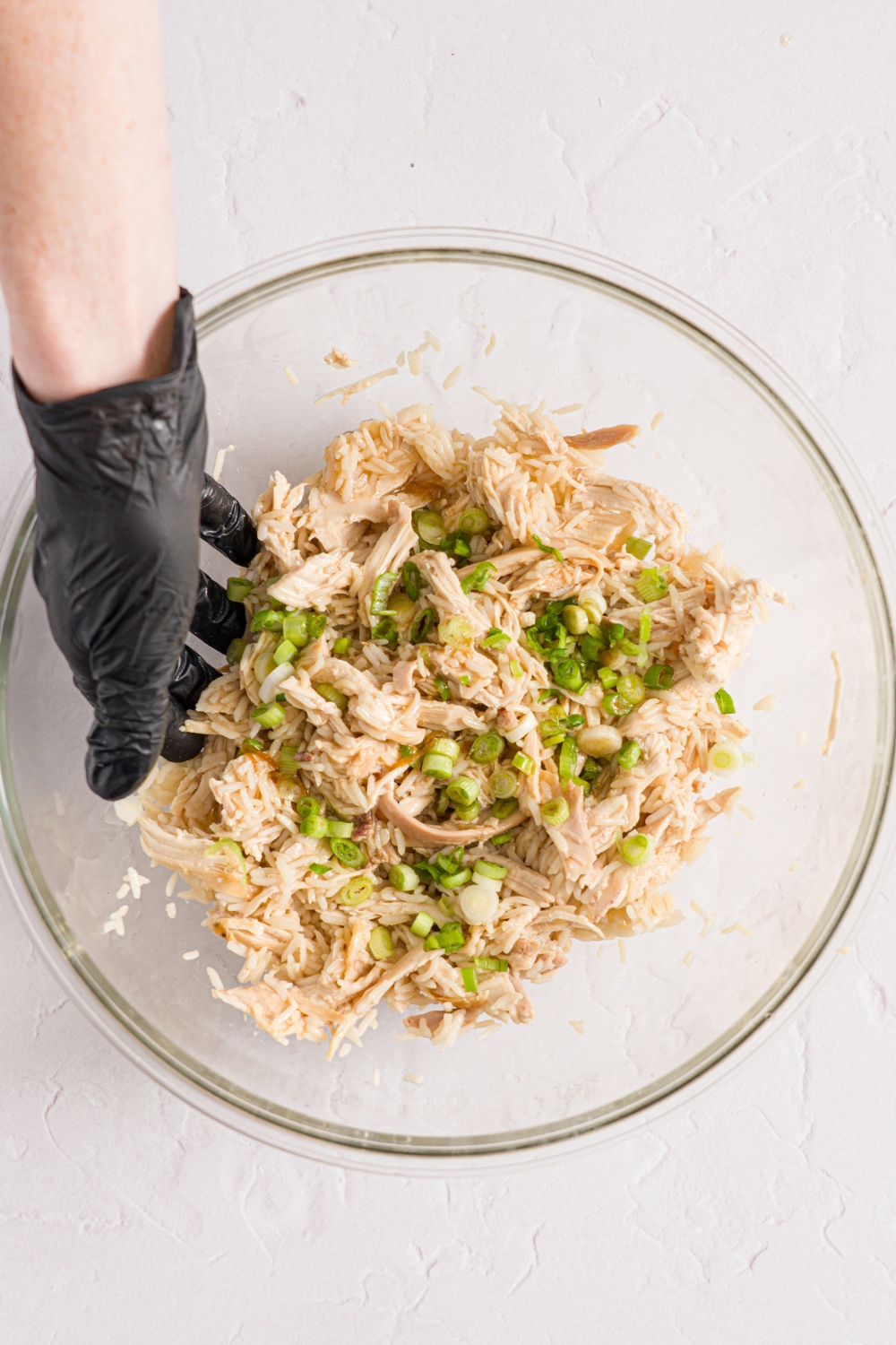 A glass bowl with a hand mixing a chicken, scallion, and rice mixture. The bowl is on a white counter.