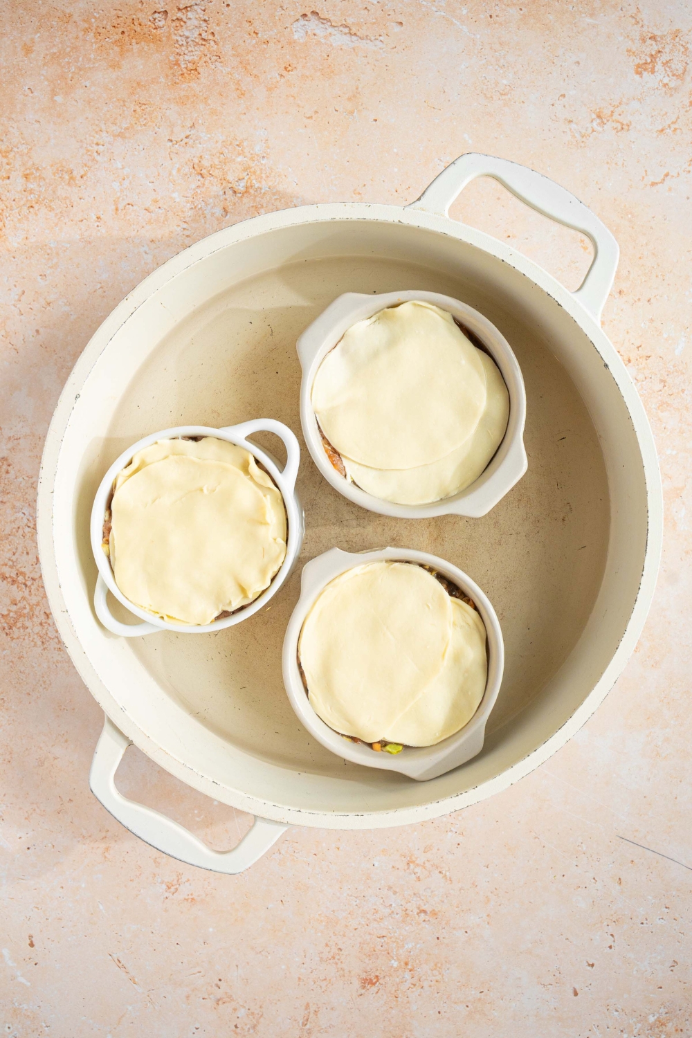 A pot with water with three ramekins with uncooked dumpling lasagna. The pot is on a tan counter.