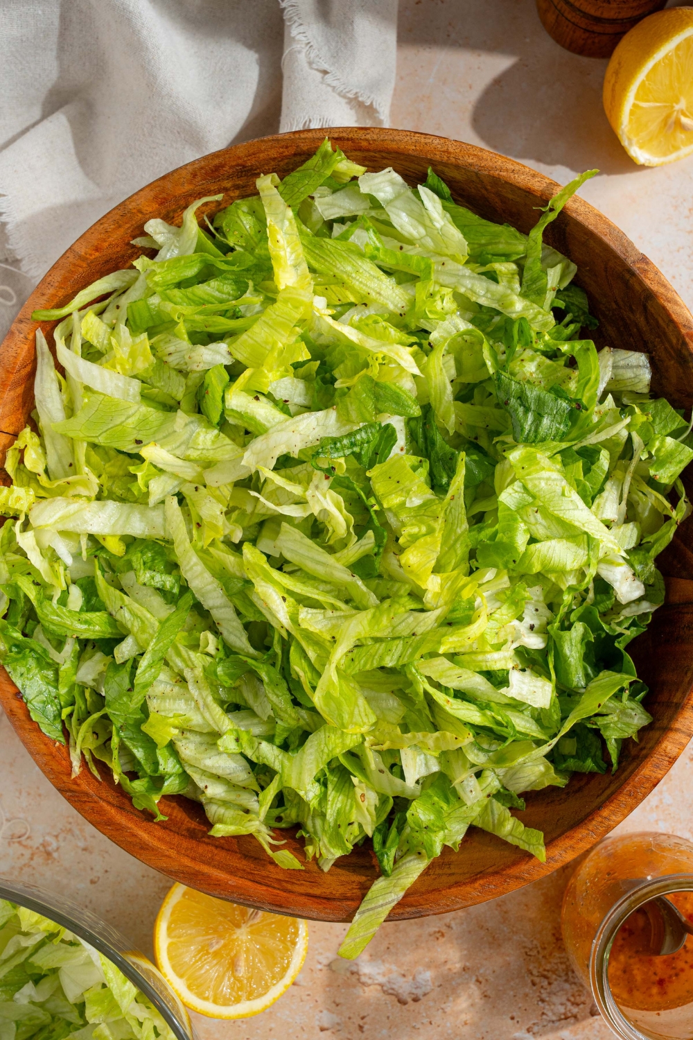 A wooden salad bowl with iceberg lettuce salad tossed in lemon dressing. The bowl is on a tan counter with sliced lemon and a white cloth napkin.