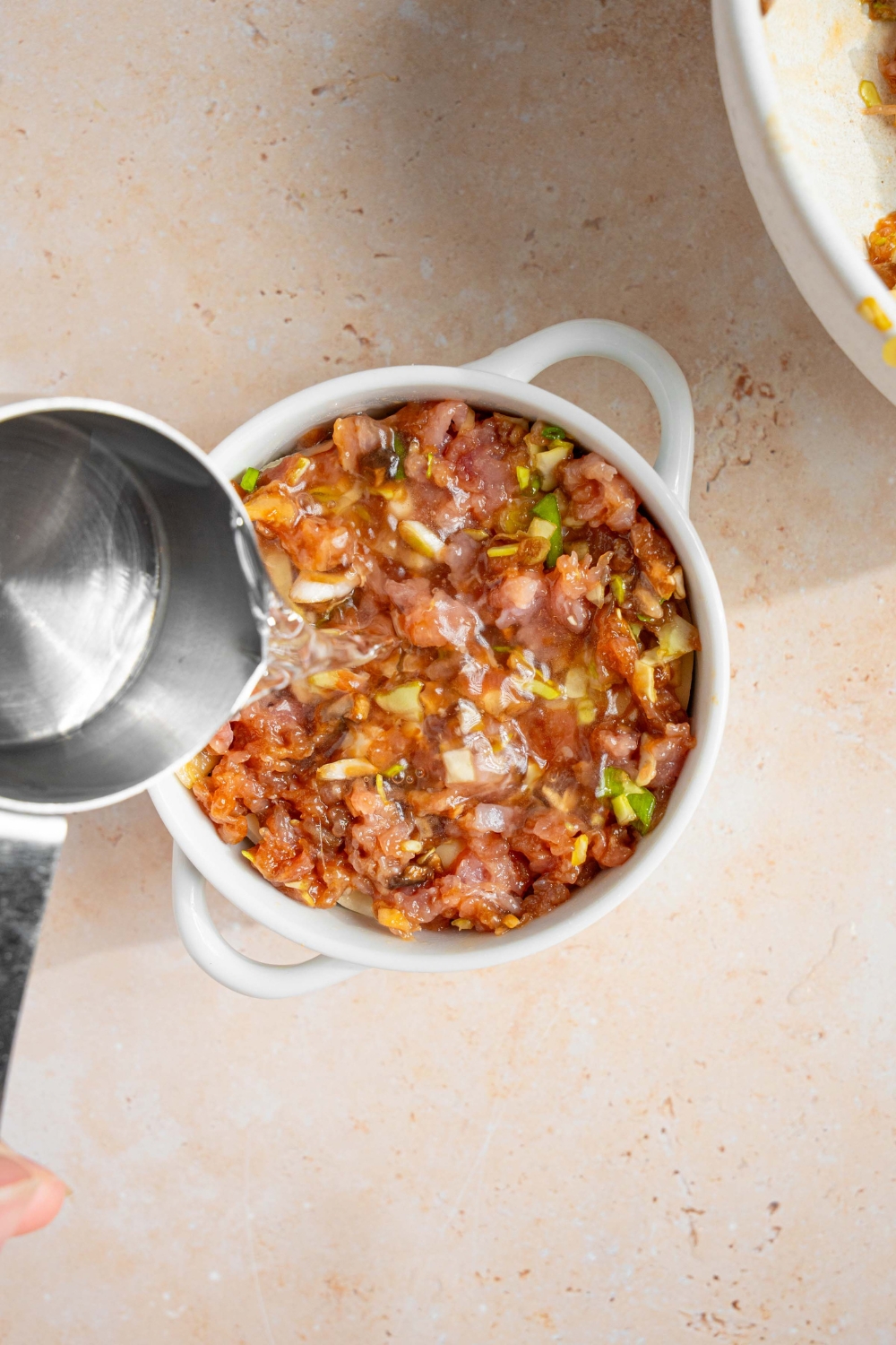 A ramekin of uncooked dumpling lasagna with the pork layer on top. A small cup is pouring water into the ramekin. The ramekin is on a tan counter.